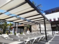 Toldo romano blanco sobre una pérgola oscura encima de tumbonas de piscina en un día soleado