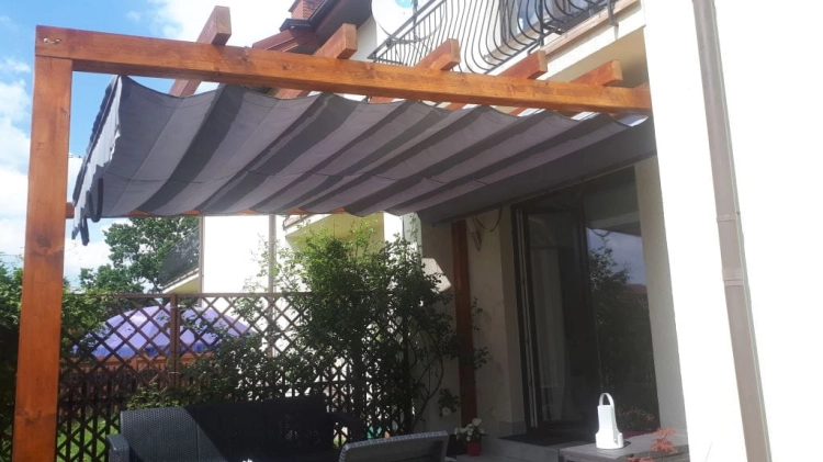 Store romain gris de terrasse 3x4 m monté sur une pergola en bois près du bâtiment, protégeant la terrasse du soleil