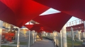 Toldo vela triangular rojo 3,2x3,2x4,5 m que crea una protección solar sobre un camino