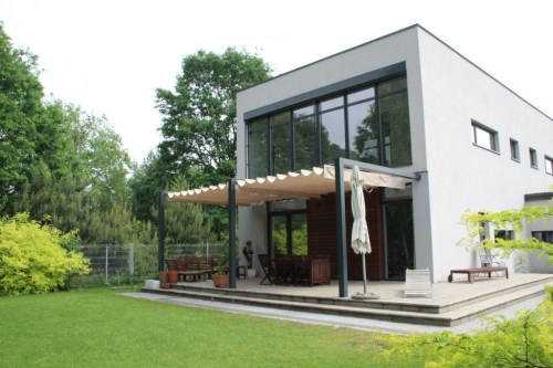 Store romain de terrasse imperméable Premium 4x4 m monté sur une structure moderne près d'une maison avec jardin