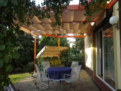Store romain de terrasse beige 4x4 m monté sur une pergola en bois au-dessus d'un salon de jardin