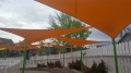 Toldos vela triangulares naranjas suspendidos sobre un parque infantil con valla blanca y postes verdes