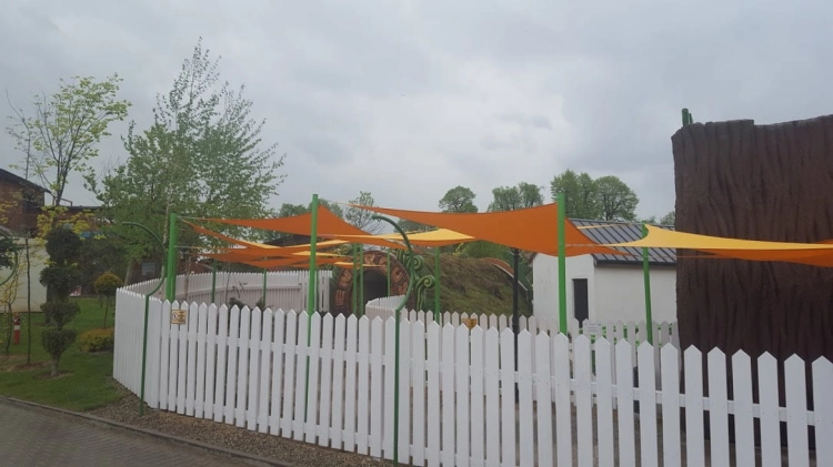 Toldo vela triangular naranja impermeable 5x5x7 m sobre parque infantil con valla blanca