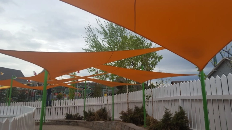 Toldo vela triangular naranja Premium instalado sobre un parque infantil junto a una valla blanca