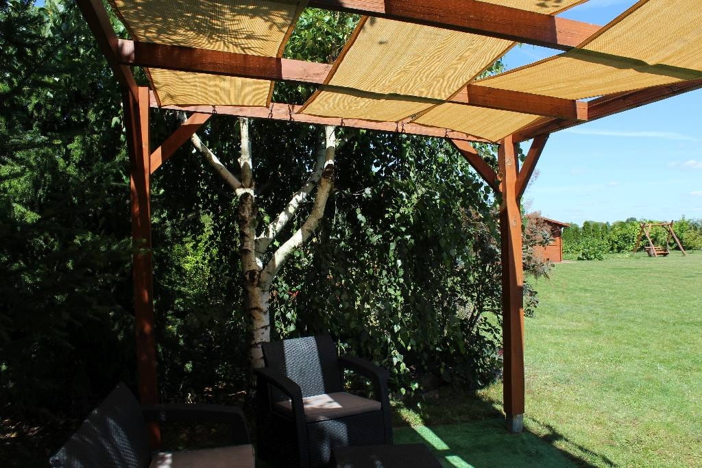 Toldo tipo banda color arena de tejido HDPE montado en una pérgola de madera en un jardín