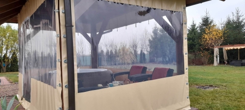 Przezroczysta plandeka na taras z beżowym obramowaniem, zamontowana na drewnianej altanie z meblami wypoczynkowymi