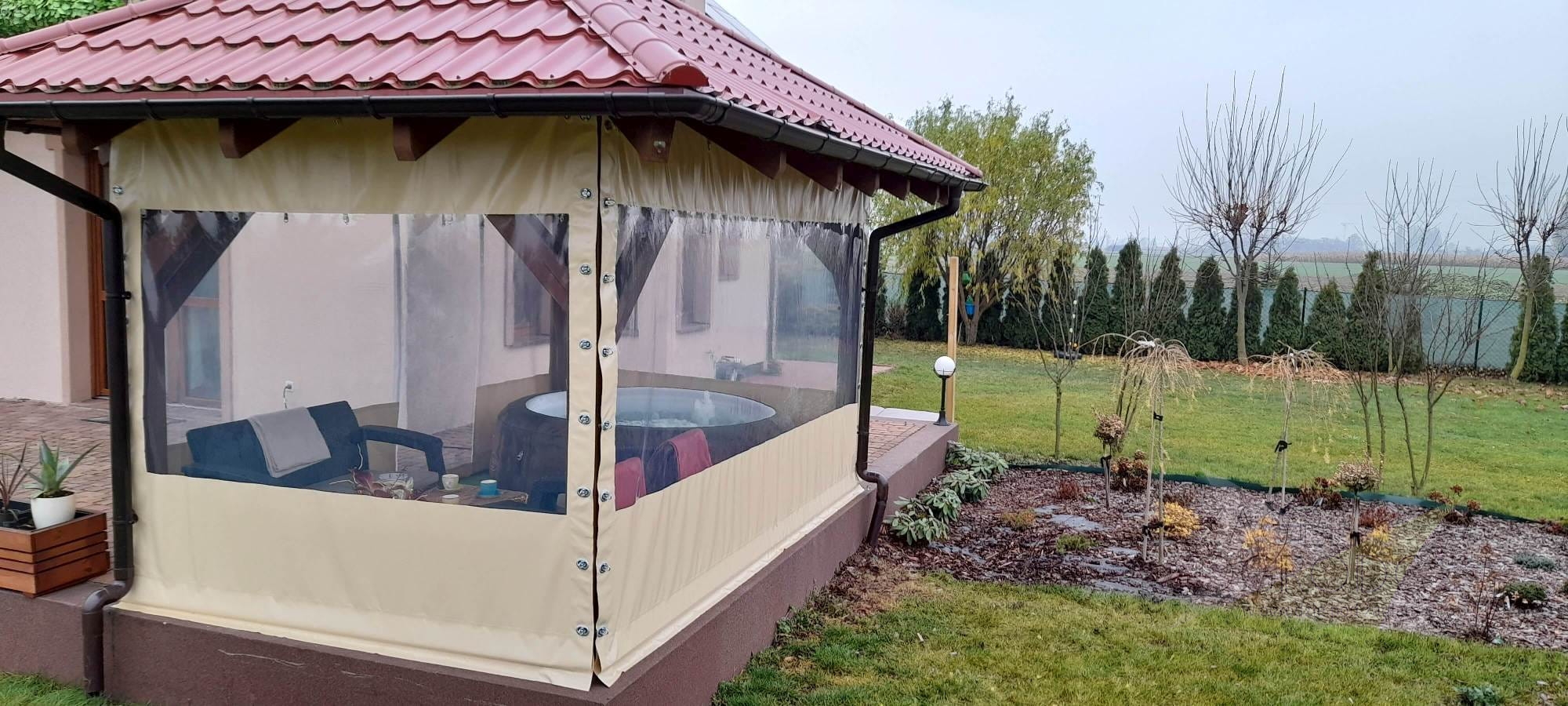 Przezroczysta plandeka na taras z beżowym obramowaniem, zamontowana na drewnianej konstrukcji altany z meblami i jacuzzi