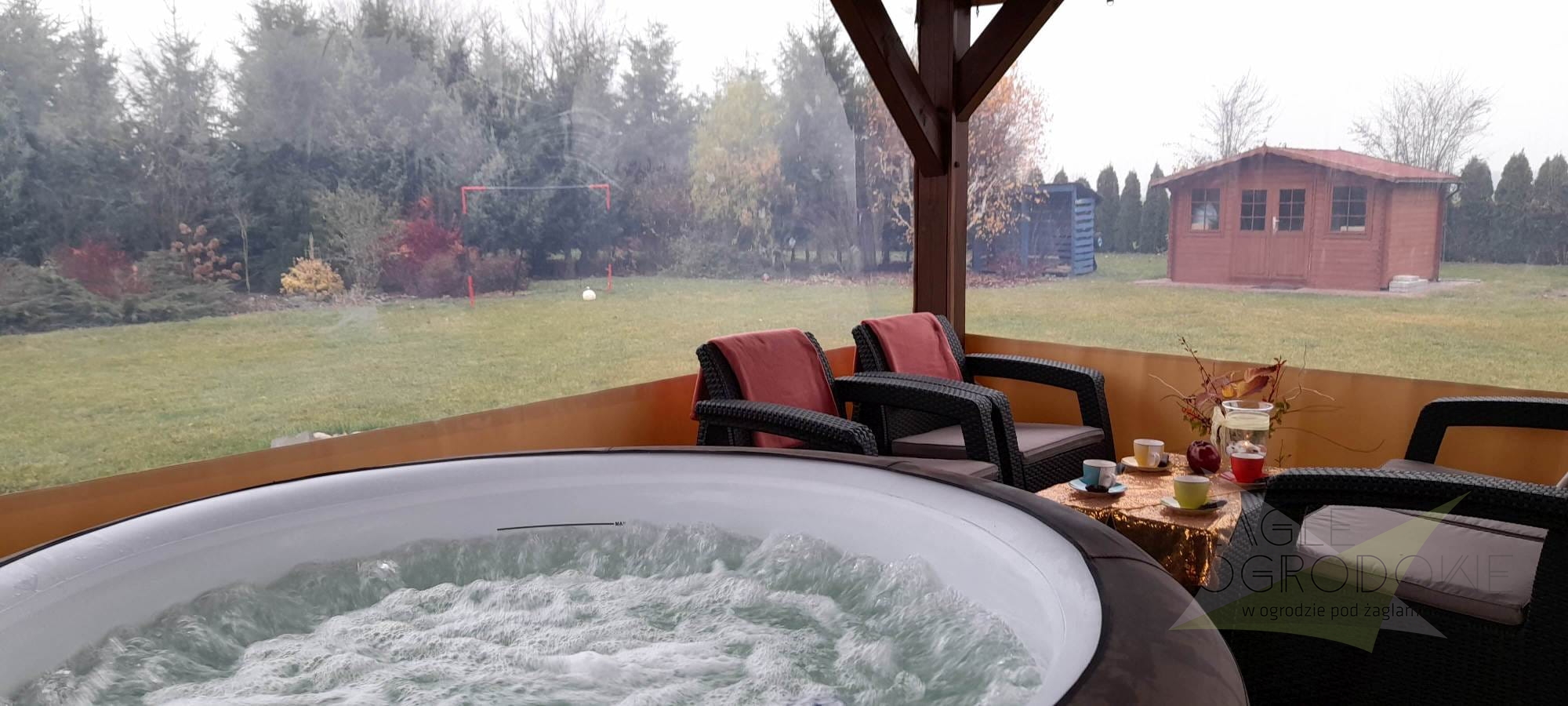 Przeźroczysta plandeka na wymiar osłaniająca taras z widokiem na ogród, przy którym stoi jacuzzi i meble ogrodowe