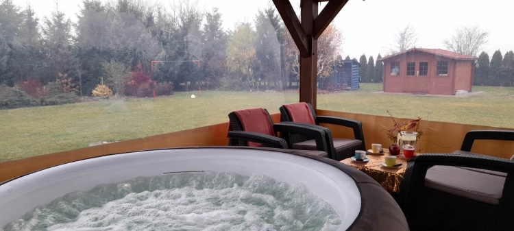 Przeźroczysta plandeka na wymiar osłaniająca taras z widokiem na ogród, przy którym stoi jacuzzi i meble ogrodowe
