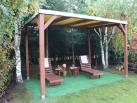 Pergola en bois avec toit en PVC beige, deux chaises longues et tables sur gazon dans un jardin