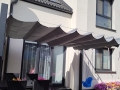 Rulou roman Premium gri pentru terasă montat deasupra unui set de mobilier de grădină pe un patio lângă o fațadă deschisă