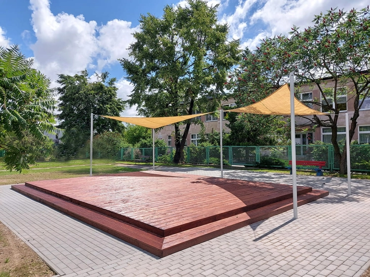 Beżowy trójkątny żagiel przeciwsłoneczny HDPE 5x5x7 m nad drewnianym tarasem w ogrodzie miejskim