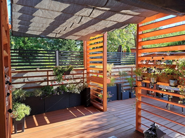 Store romain pare-soleil gris en HDPE monté sur une terrasse en bois avec pergola et plantes en pots