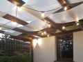 Wit zonnezeil gevlochten tussen de houten balken van een pergola met lichtsnoer