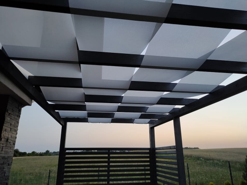 Voile d'ombrage gris clair 1x5 m entrelacée entre les poutres d'une pergola de jardin sombre sur fond de champ