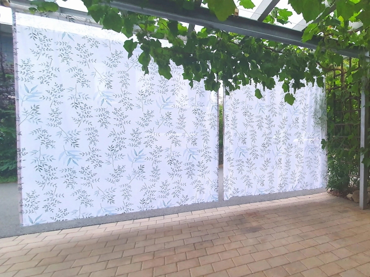 Rideau de jardin blanc avec imprimé feuilles vertes et bleues, suspendu sur une terrasse sous une pergola végétalisée