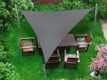 Toldo vela triangular gris sobre muebles de jardín de madera en un césped rodeado de vegetación