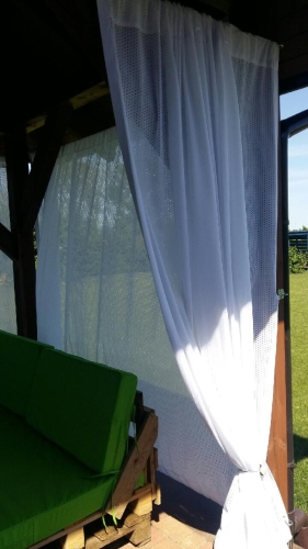 Cortina blanca de terraza colgada en un cenador de madera junto a un sofá de jardín verde de palets