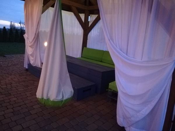 Cortina de terraza tipo visillo blanca decorando un cenador de madera con muebles de jardín verdes a medida