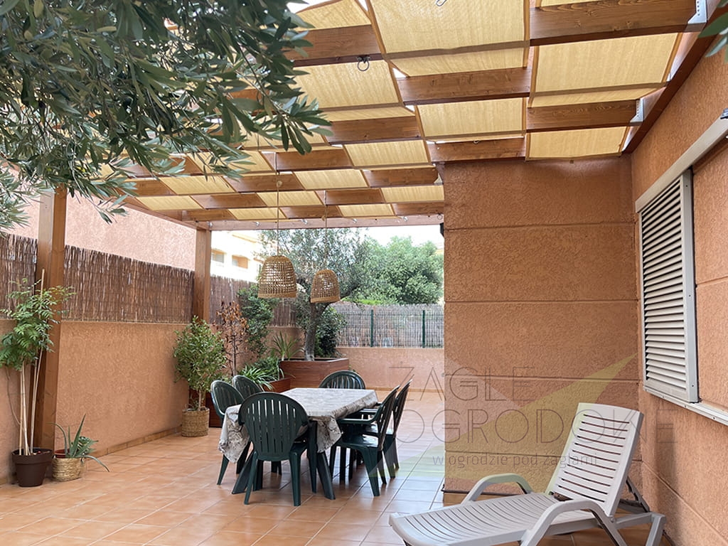 Toldo tipo banda beige de tejido HDPE montado sobre pérgola de madera en terraza con muebles de jardín