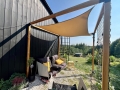 Zandkleurig schaduwdoek op maat gemonteerd boven terrasmeubels bij een houten gevel