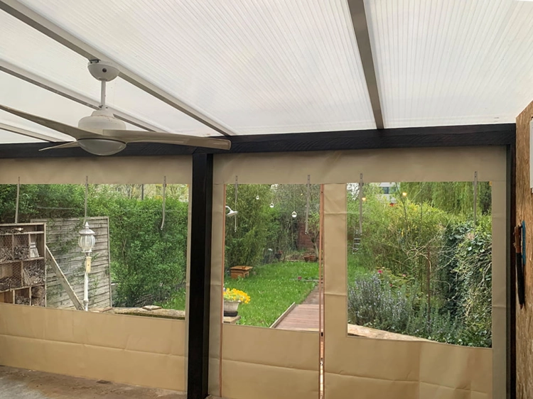 Transparant dekzeil voor terras met beige rand en raam, gemonteerd onder een overkapping met ventilator