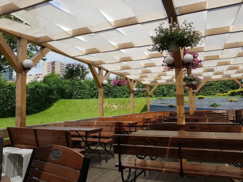 Wit zonneschermdoek gemonteerd op een houten pergola in een restauranttuin met banken en bloemen