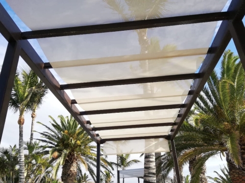 Wit Romeins zonnezeil gemonteerd op een bruine buitenpergola met uitzicht op palmbomen