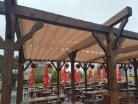 Store romain de terrasse Premium imperméable beige, monté sur une pergola en bois au-dessus des tables