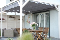 Graues Raffrollo, sonnenfest und wasserdicht, montiert an einer weißen Holzpergola über der Terrasse mit Möbeln