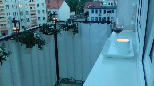 Biała, matowa osłona balkonowa na wymiar zamontowana na balustradzie z kwiatami, obok kieliszek wina i świeca