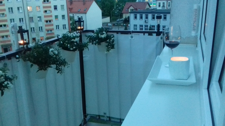 Biała, matowa osłona balkonowa na wymiar zamontowana na balustradzie z kwiatami, obok kieliszek wina i świeca