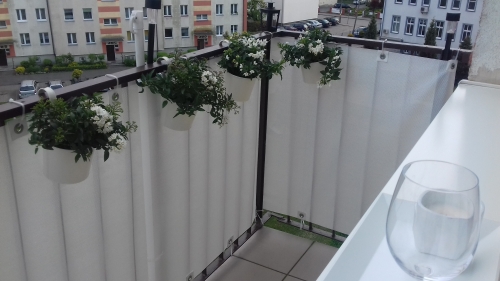 Biała mata osłonowa na poręczy balkonu z wiszącymi doniczkami z białymi kwiatami na tle osiedla mieszkaniowego