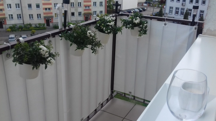 Biała mata osłonowa na wymiar zamontowana na balustradzie balkonu z białymi doniczkami z kwiatami