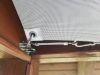 Fijación de un toldo vela retráctil blanco con mosquetón, tensor y soporte en una viga de madera