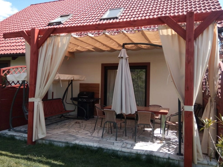 Biela záhradná clona vodotesná na mieru namontovaná v drevenej pergole na terase s relaxačným nábytkom